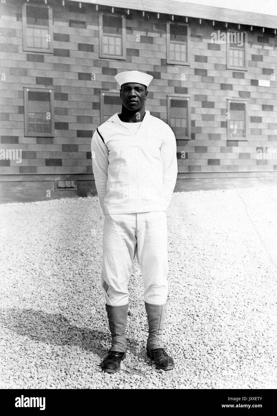 Ritratto di un afro-americano us navy sailor in piedi nella parte anteriore dell'edificio, le sue mani sono dietro la schiena e indossa una luce colorata uniforme del marinaio, 1920. Foto Stock
