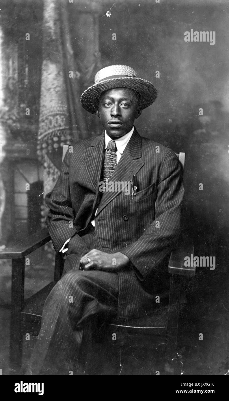 Ritratto a lunghezza intera di uomo afroamericano maturo, con vestito a righe scure, cravatta con design e cappello, penne in tasca della giacca, seduta sulla sedia davanti allo sfondo, espressione neutra, 1925. Foto Stock