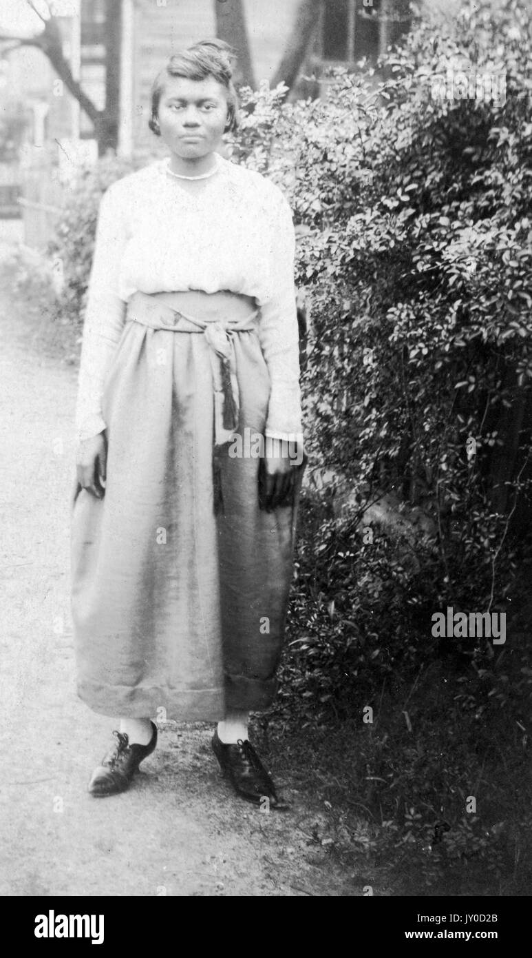 Ritratto di una donna afro-americana in piedi accanto a cespugli alti, lei indossa una lunga gonna colorata luce e una blusa colorata luce, lei indossa una collana, le sue mani sono ai suoi lati, 1915. Foto Stock