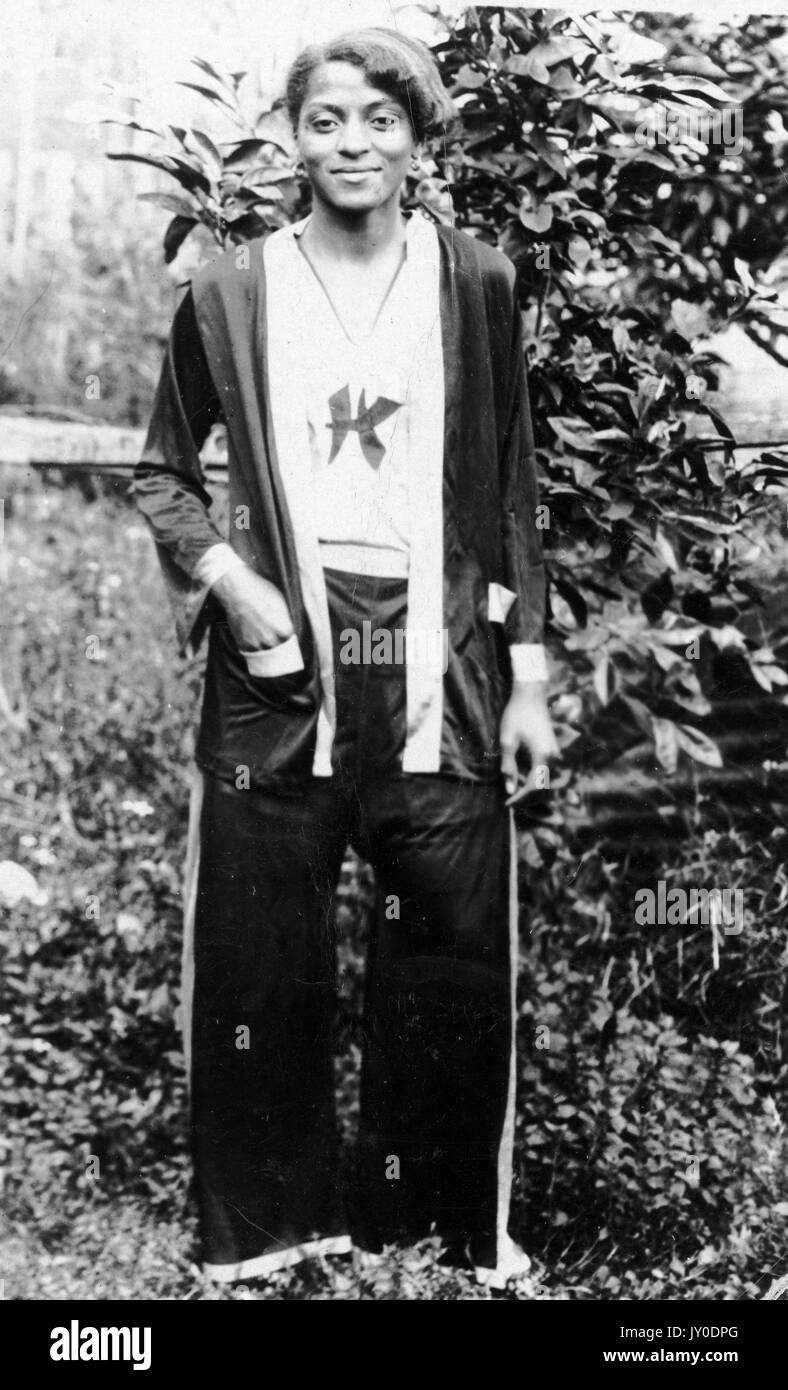 Ritratto di una donna afro-americana sorridente e in piedi davanti a una cespupa, indossa pantaloni lunghi e scuri e un pullover abbinato, indossa una camicia di colore chiaro sotto, una mano è in tasca e l'altra al suo fianco, 1929. Foto Stock