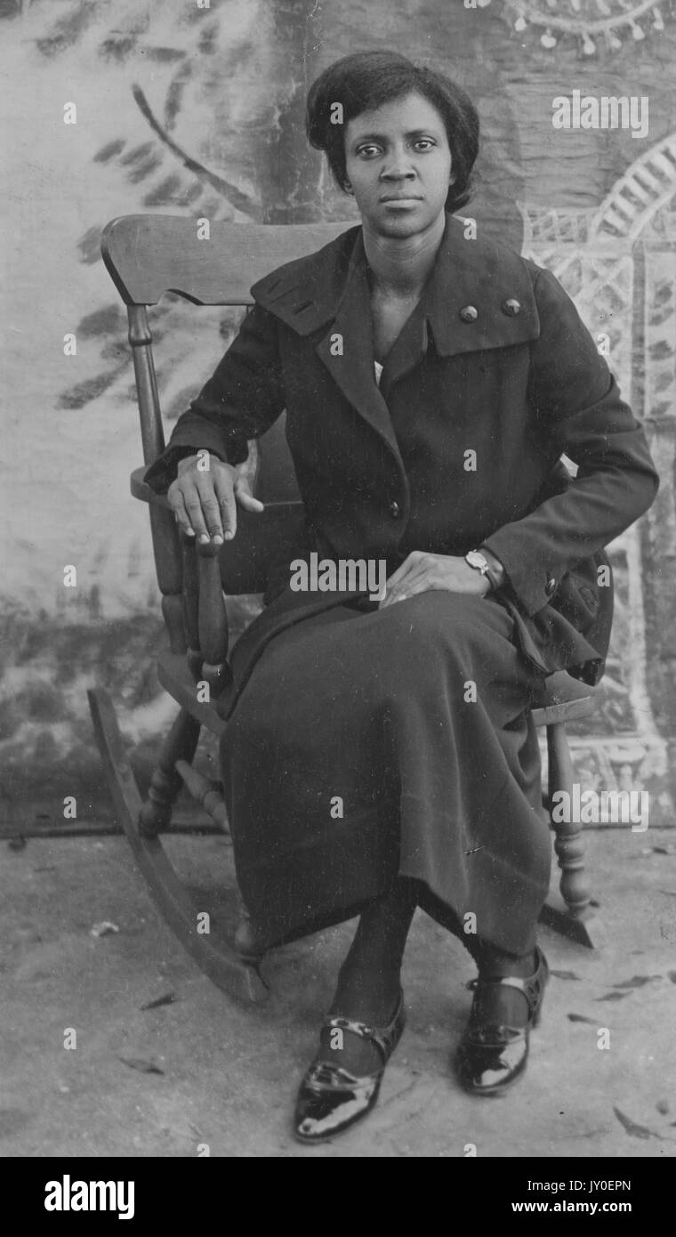 Ritratto a lunghezza intera di donna afroamericana matura con espressione neutra davanti ad uno sfondo dipinto, seduto su una sedia basculante in legno, indossando un cappotto scuro, gonna, e scarpe, 1925. Foto Stock