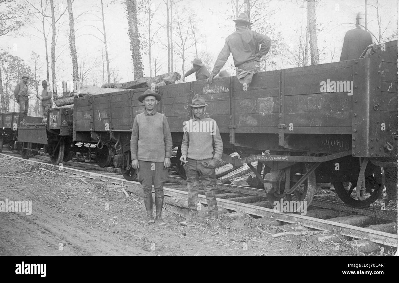 Due uomini si trovano all'esterno in una zona boscosa, di fronte ai treni con le vetture da boxcar, su cui sono in piedi altri lavoratori, 1920. Foto Stock