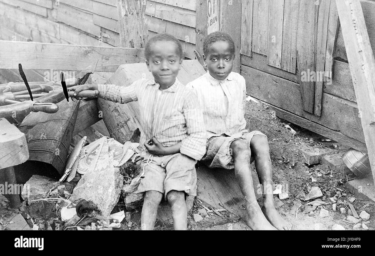 Ritratto a lunghezza intera di due bambini afro-americani, con magliette e shorts a righe, seduti in un mucchio di rifiuti e sporcizia davanti al vecchio magazzino, espressioni neutre, 1920. Foto Stock