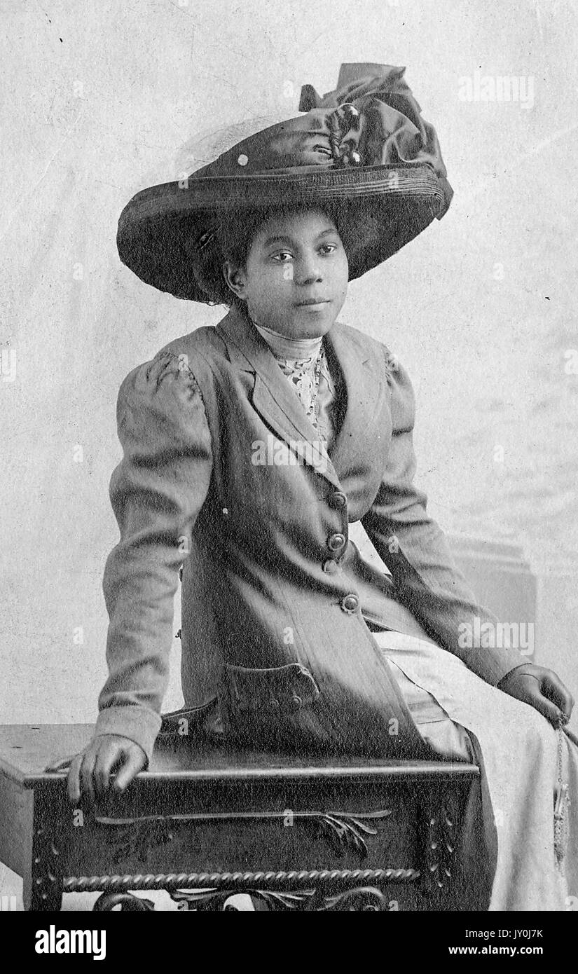 Ritratto di una giovane donna afro-americana che sting su un piccolo tavolo di legno, sta portando un vestito di colore chiaro e un cappotto di fondo di colore chiaro sulla parte superiore, sta portando un grande cappello decorato, 1915. Foto Stock