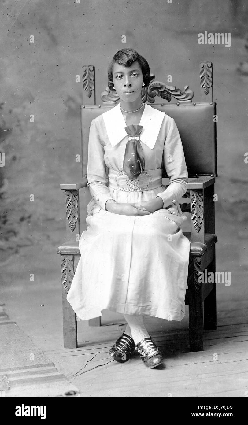 Ritratto di un americano africano donna seduta in un grande in legno intagliato sedia, indossa una luce lungo mantello colorato e una luce superiore colorato che viene infilato in essa è anche indossando un grande tirante e orecchini perla, le sue mani sono appoggiate in grembo, 1915. Foto Stock
