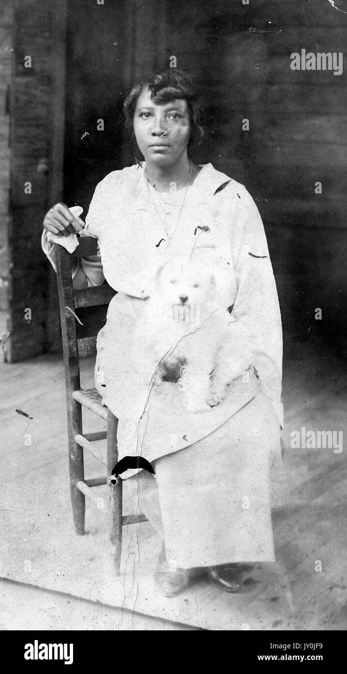 Ritratto di una donna afro-americana seduta in una sedia di legno, sta indossando una gonna e gonna di colore chiaro, c'è un cane di colore chiaro seduto in grembo, il suo braccio destro è sulla schiena della sedia e il suo braccio sinistro è dal cane, 1915. Foto Stock