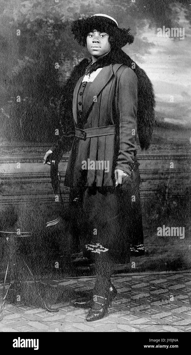 Ritratto di una donna afro-americana che si trova di fronte ad un muro con un murale, il suo braccio destro è posto su una sedia a sinistra e il suo braccio sinistro è al suo fianco, lei indossa una gonna di colore scuro e un cappotto di colore scuro, indossa una stola in pelliccia e un cappello, 1915. Foto Stock
