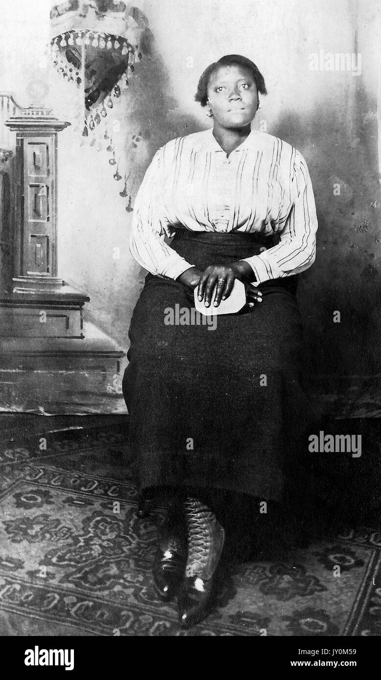 Ritratto a tutta lunghezza di donna afro-americana, seduta davanti a uno sfondo, con le mani piegate in grembo, con un abito scuro e una blusa a righe, con una seria espressione facciale, 1920. Foto Stock