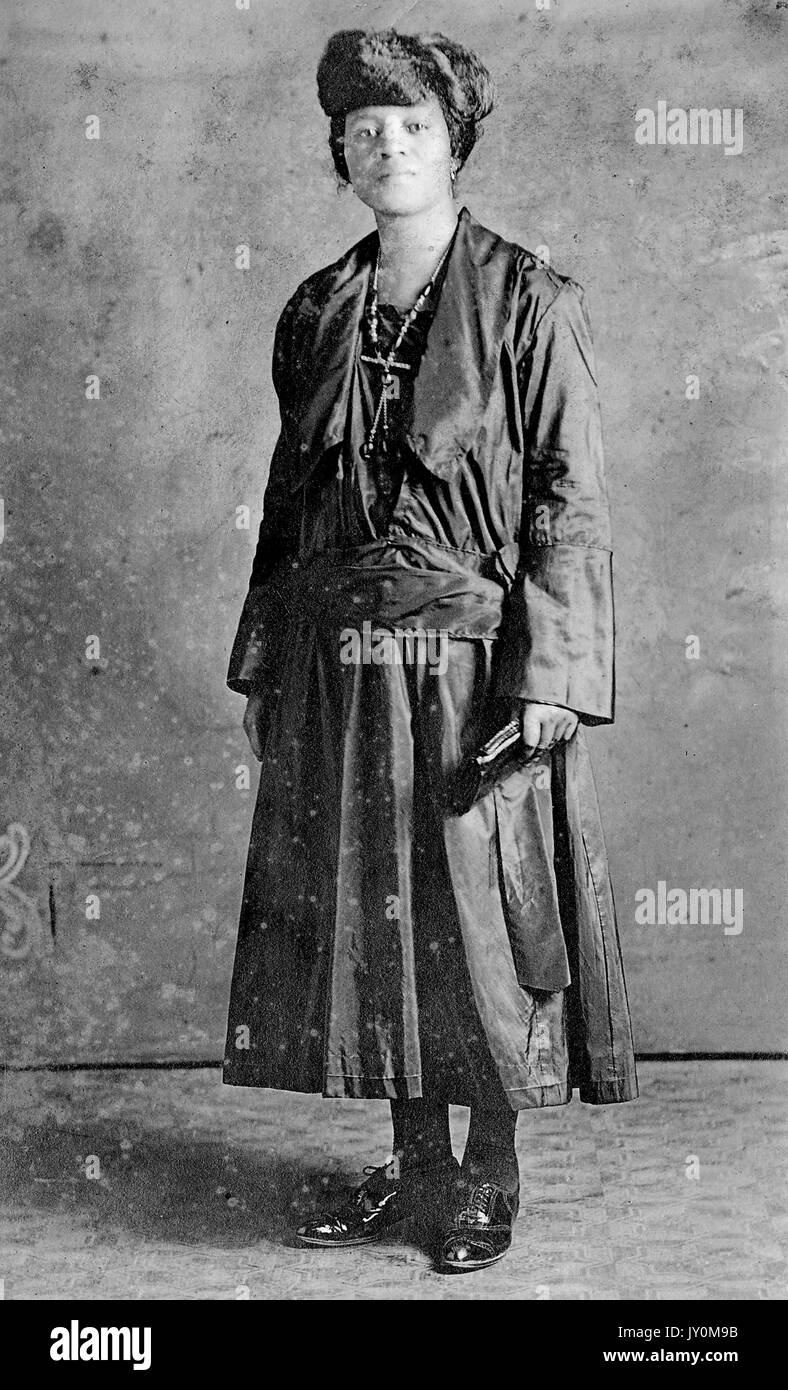 Ritratto a figura intera di African American donna in piedi di fronte a uno sfondo, in un abito nero e nero scarpe, stringendo un portamonete, entrambi i bracci dal suo lato, con un grave espressione facciale, 1920. Foto Stock