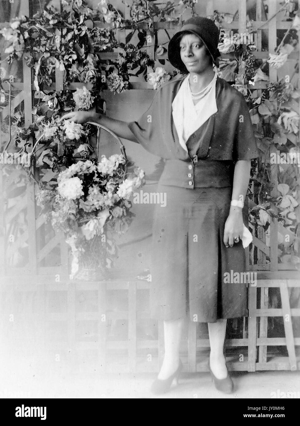 Ritratto a corpo pieno di donna afroamericana in piedi davanti ad una recinzione adornata con fiori bianchi, come lei tiene un cesto dei fiori nella sua mano destra, indossando un abito scuro, tenendo un fazzoletto nella sua altra mano, con una seria espressione facciale, 1920. Foto Stock