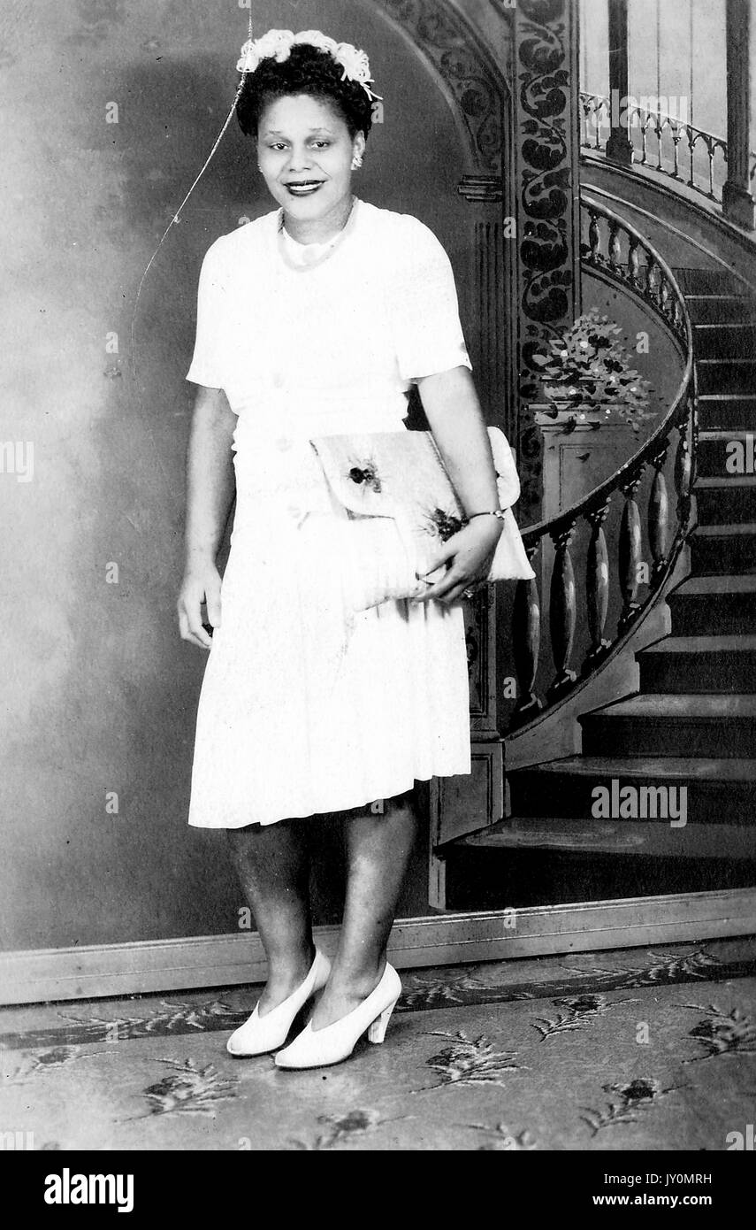 Ritratto a corpo pieno di donna afroamericana, indossando un abito bianco e scarpe bianche, stringendo la sua borsa, in piedi davanti a uno sfondo di una scala, con un'espressione sorridente del viso, 1935. Foto Stock