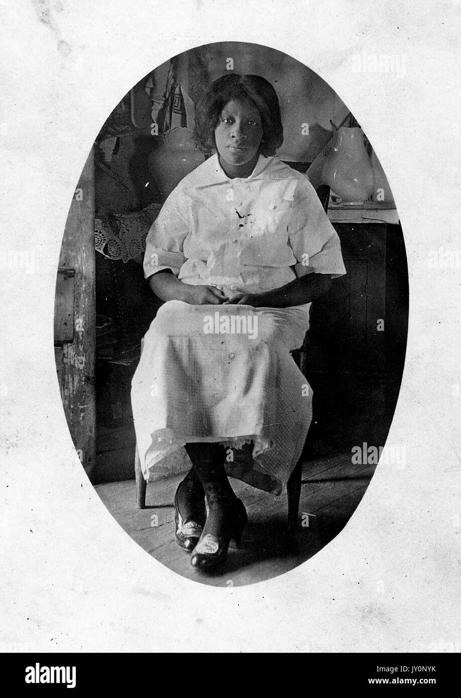 Ritratto seduta a lunghezza intera, donna afroamericana matura, vestita leggera, seduta davanti al mobile con vasi, espressione neutra, 1915. Foto Stock