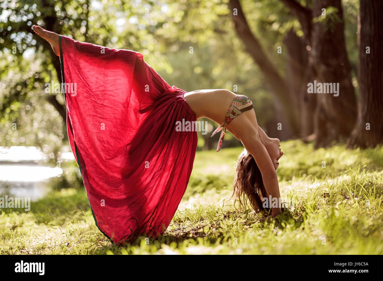 Montare il giovane bella donna che indossa il mantello rosso che lavora fuori all'aperto nel parco sulla giornata estiva, facendo ponte pongono da yoga Foto Stock
