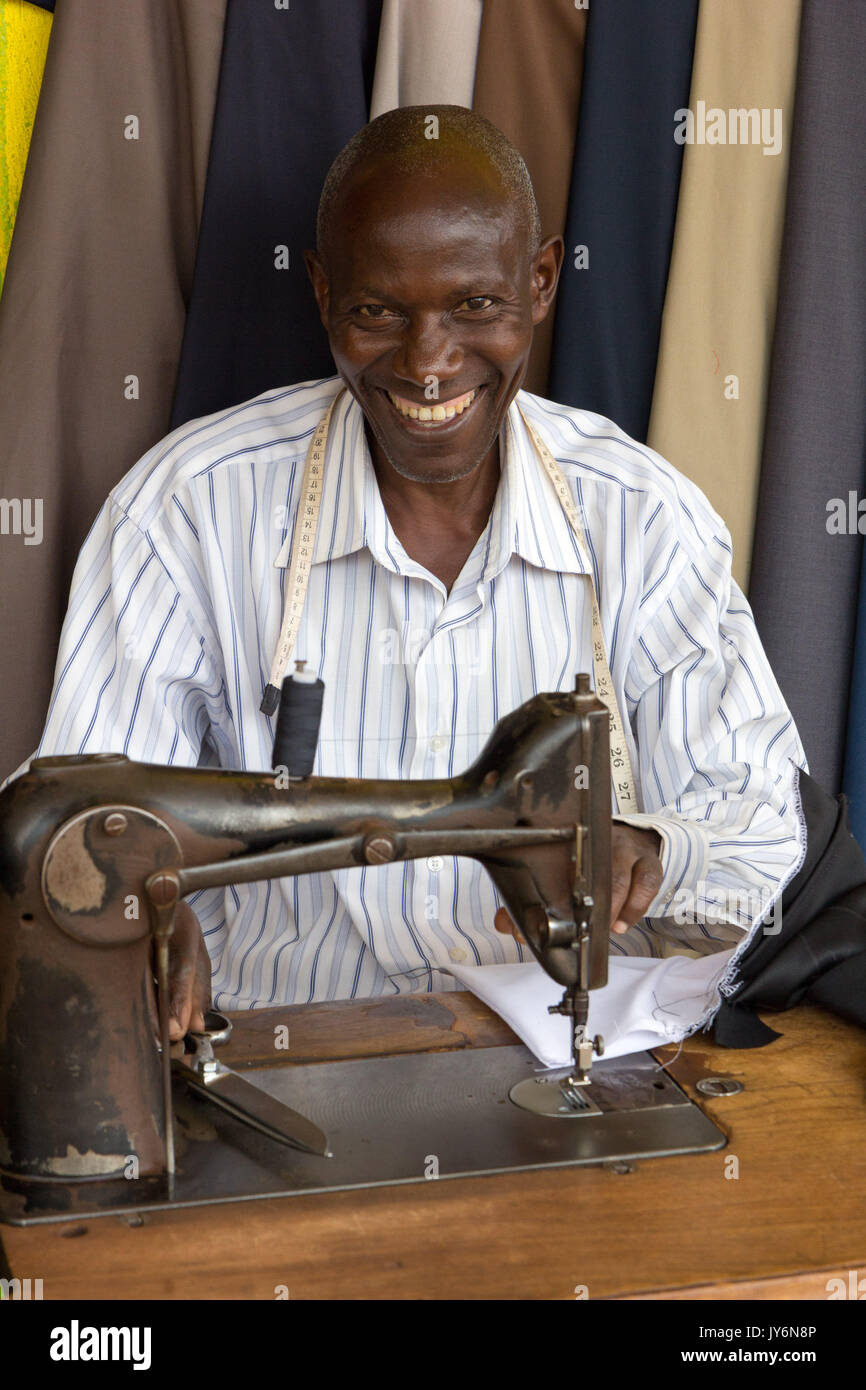 Un sorridente couturier africani presso la sua macchina per cucire in un salotto di strada lavorando su alcuni vestiti. Foto Stock