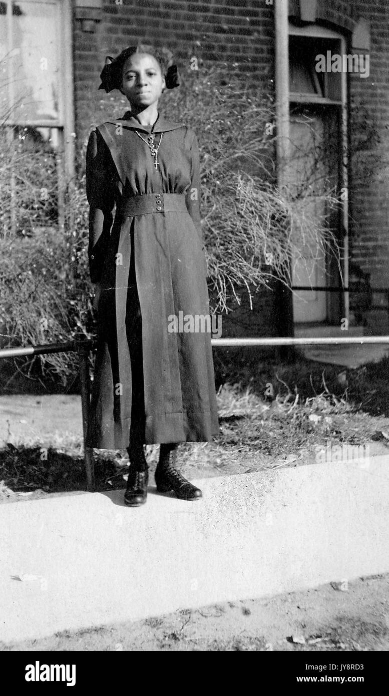 A piena lunghezza ritratto permanente del giovane africano donna americana, indossa un abito scuro, una collana e un'espressione neutra, in piedi su un percorso nella parte anteriore di piante e di un edificio, 1915. Foto Stock