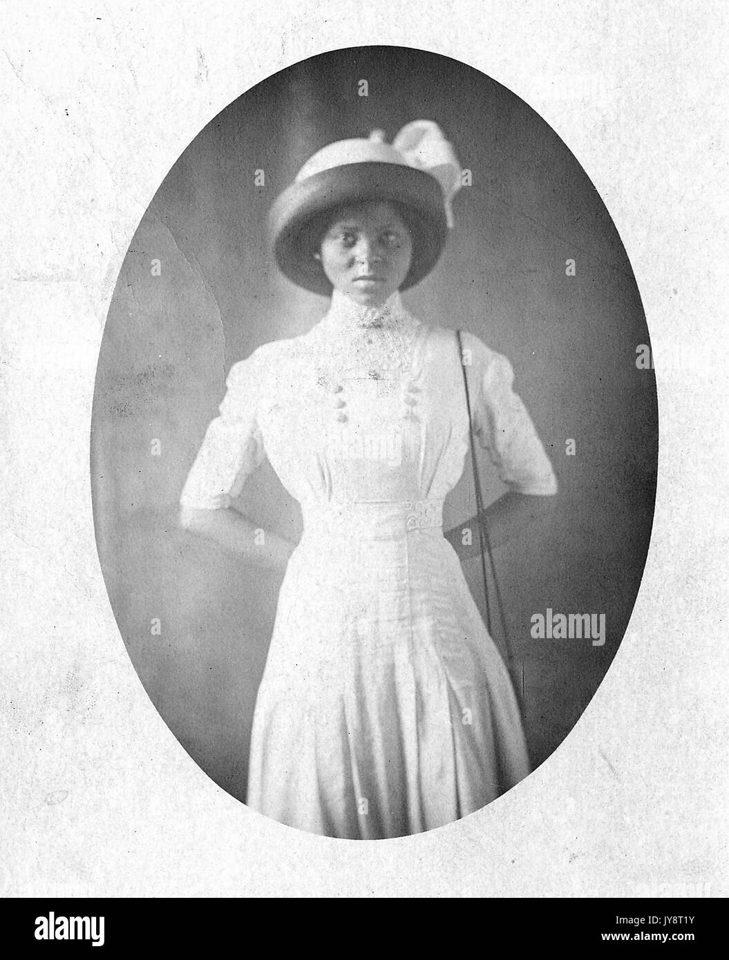 Tre quarti lunghezza Ritratto di giovane americano africano donna con cappello formale e abito bianco, con le mani incrociate dietro la schiena con un grave espressione facciale, 1915. Foto Stock