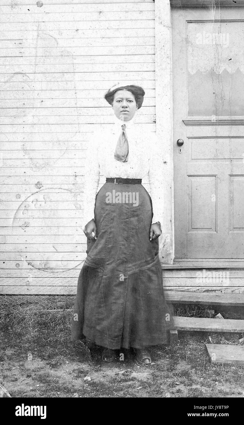 A piena lunghezza Ritratto di African American donna che indossa camicia formale con cravatta e gonna lunga, in piedi al di fuori di un edificio in legno, le braccia ai suoi lati con una seria espressione facciale, 1911. Foto Stock