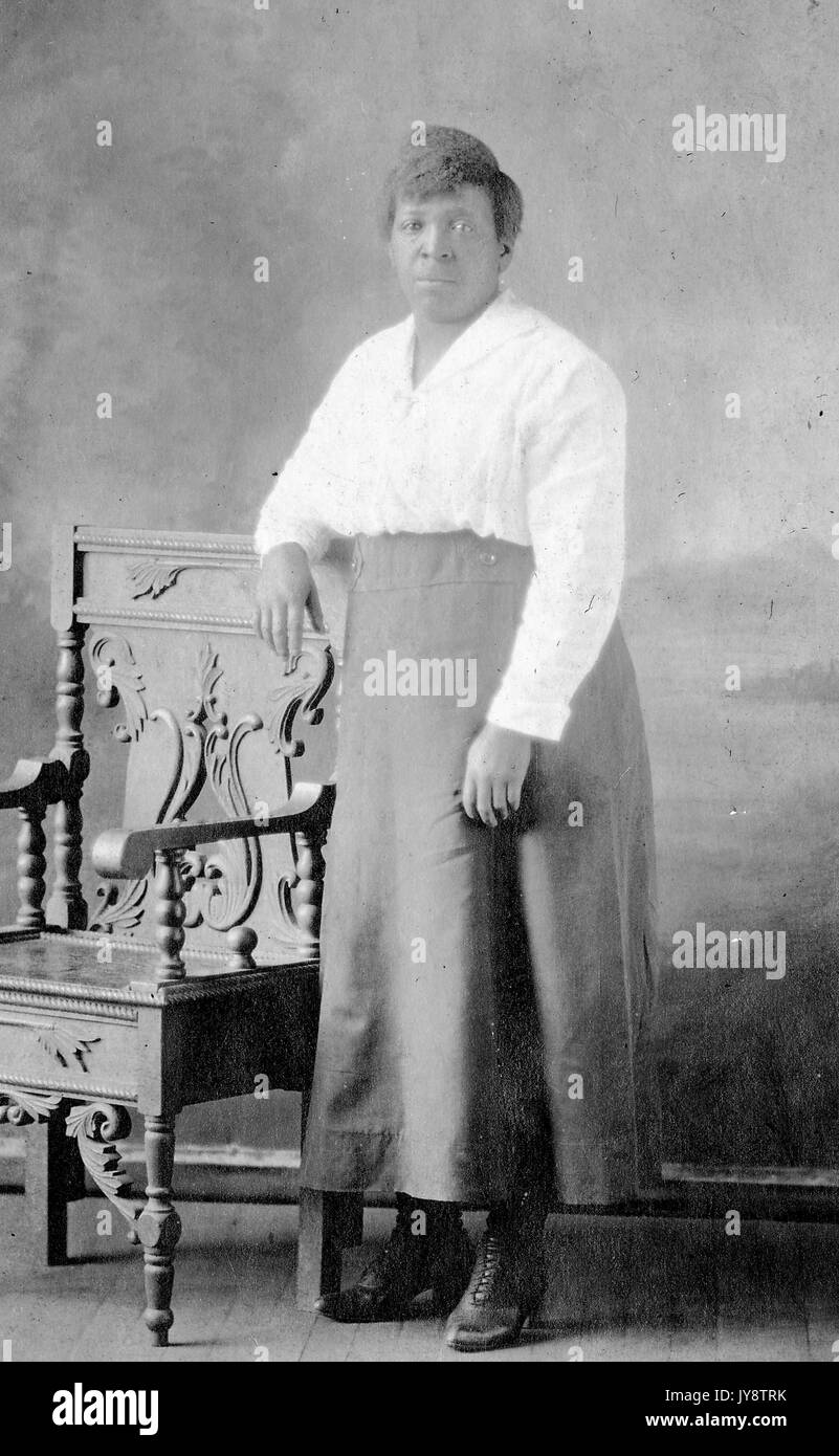 Ritratto a lunghezza intera di donna afroamericana matura in piedi con il suo braccio poggiato sulla schiena di una sedia in legno ornato, espressione facciale neutra, 1944. Foto Stock
