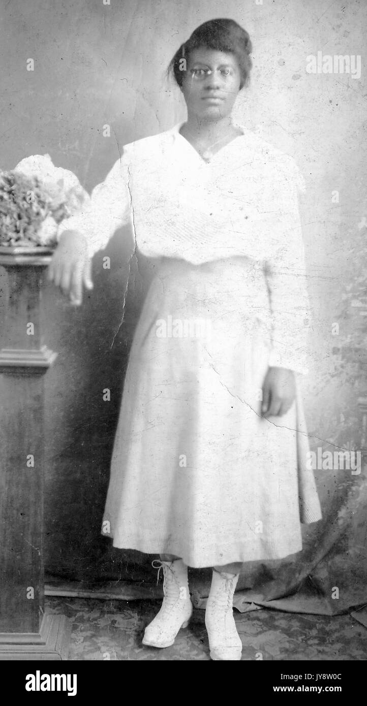 A piena lunghezza Ritratto di giovane americano africano donna che indossa gli occhiali, una camicia bianca e stivali e piedi con il suo braccio appoggiato su un tavolo, 1944. Foto Stock