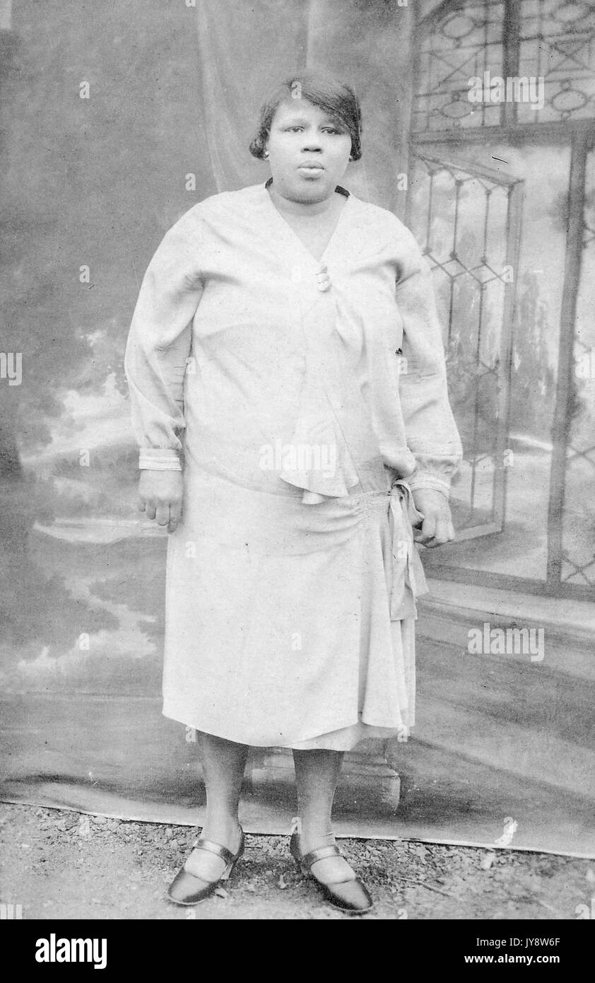 A piena lunghezza Ritratto di African American donna in piedi sul tracciato sterrato, davanti ad un fondale dipinto che indossa un abito bianco con pensieroso espressione facciale, 1930. Foto Stock