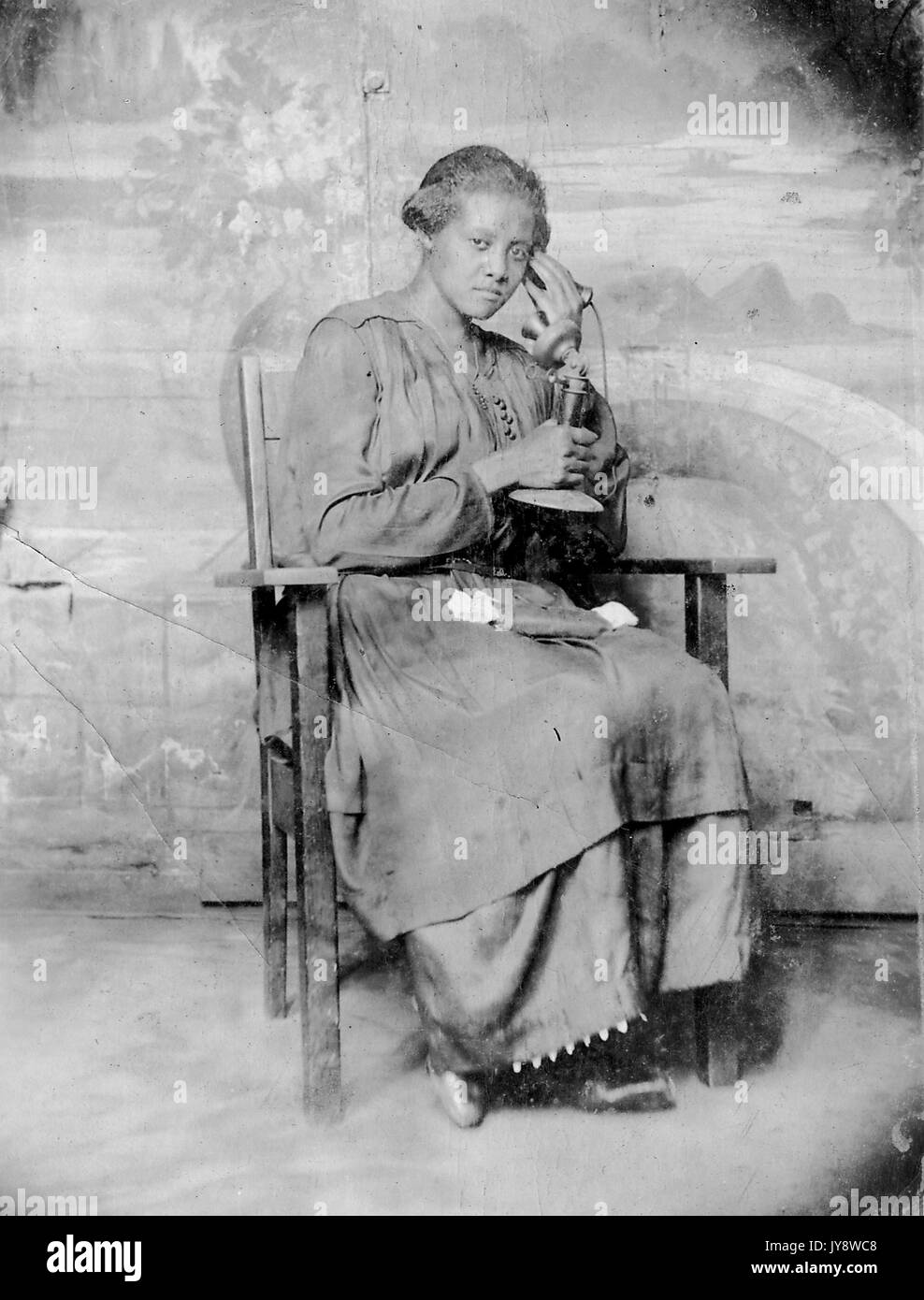 A piena lunghezza Ritratto di African American donna, seduto in una sedia in legno, indossando un abito scuro, sul telefono con un grave espressione facciale, 1920. Foto Stock