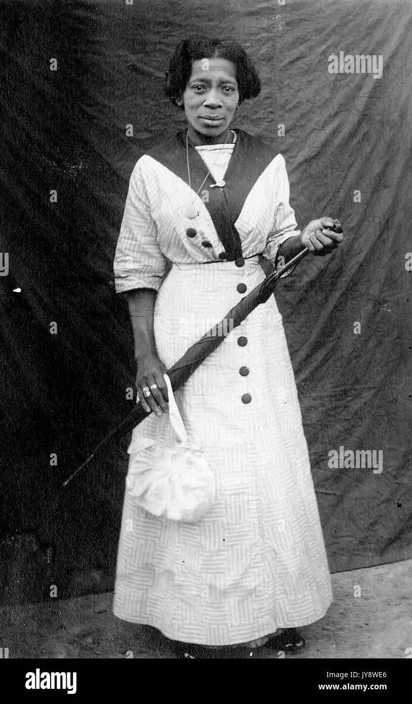 A piena lunghezza Ritratto di African American donna in piedi di fronte a uno sfondo scuro, indossando un abito bianco, tenendo un portamonete bianco e un ombrello scuro, con un grave espressione facciale, 1920. Foto Stock