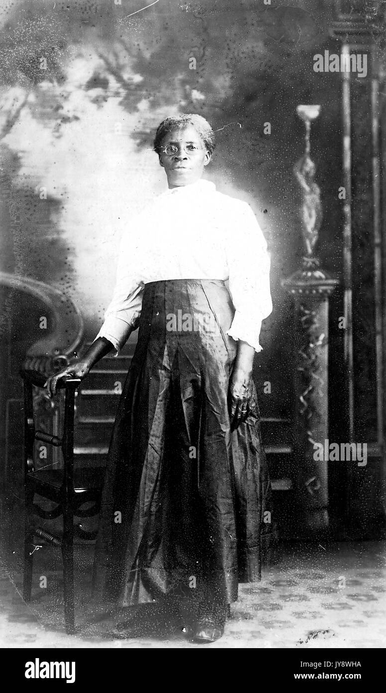 A piena lunghezza Ritratto di African American donna che indossa un abito scuro, in piedi di fronte ad una sedia in legno con la sua mano appoggiata su di essa, poste nella parte anteriore di un affresco-come sfondo, con un grave espressione facciale, 1920. Foto Stock