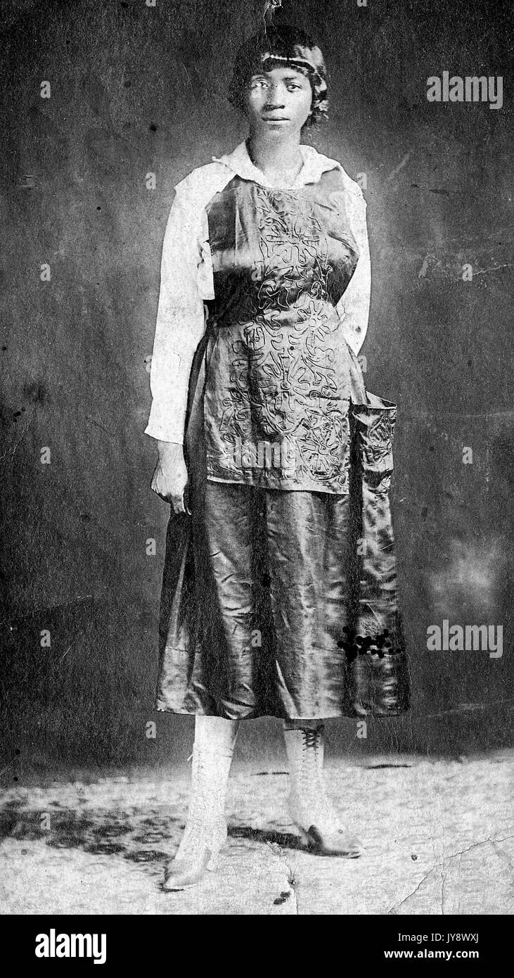 A piena lunghezza Ritratto di un africano donna americana, archly permanente, indossando un scuro abito ricamato con scarpe di bianco, con un grave espressione facciale, di fronte a uno sfondo scuro, 1920. Foto Stock