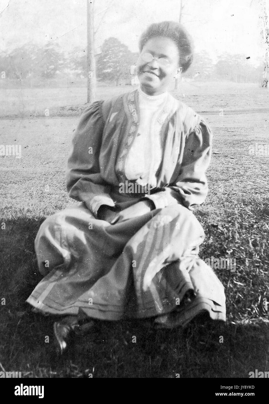 A piena lunghezza ritratto, seduti african american donna, all'aperto seduti con le gambe incrociate in erba, indossando abiti lunghi, indossando occhiali, sorridente espressione facciale, 1915. Foto Stock