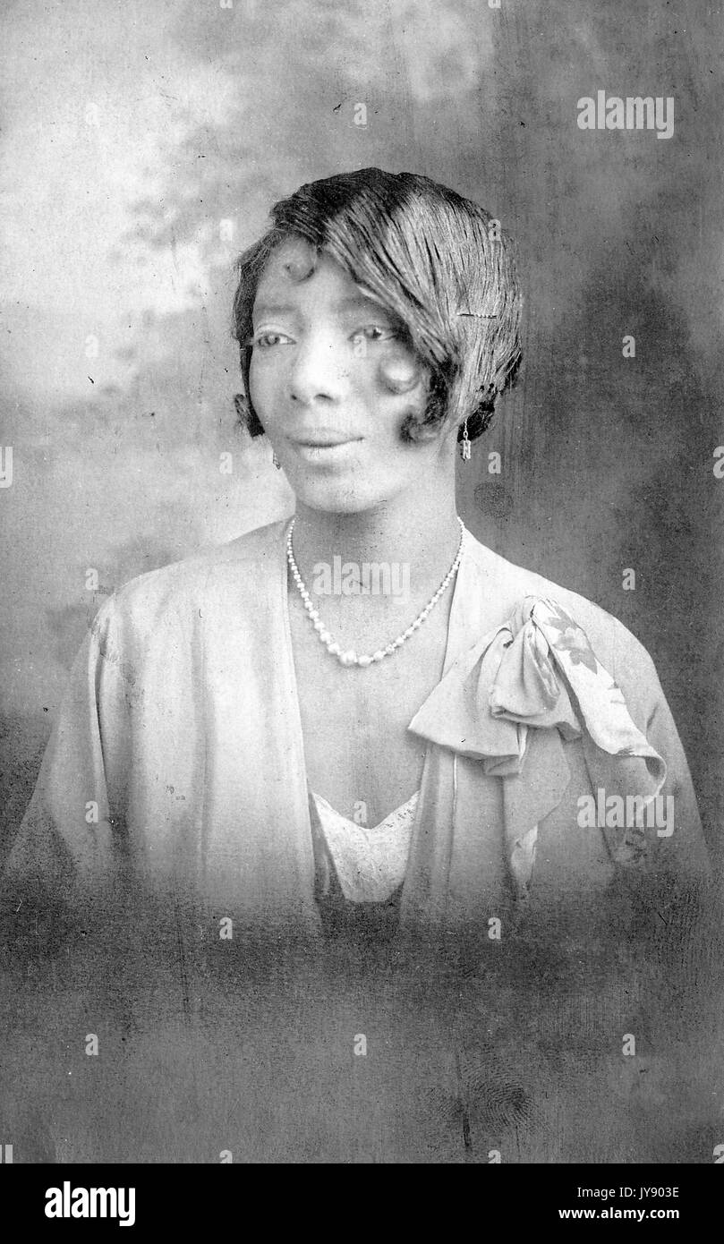 Colpo di testa di matura african donna americana, indossa una camicetta di luce con perla collana e orecchini, espressione neutra, 1925. Foto Stock