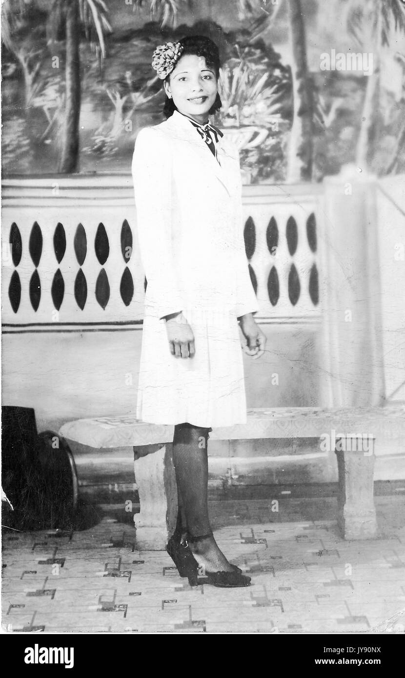 Ritratto di un americano africano donna in piedi di fronte a un banco che indossa un abito bianco con talloni e un fiore tra i capelli, 1920. Foto Stock