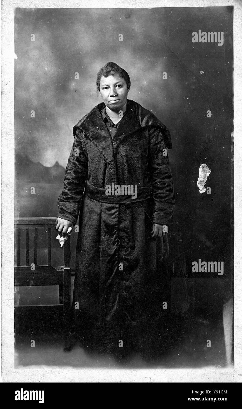 Ritratto di una donna afroamericana matura a lunghezza intera con un'espressione neutra, con un cappotto scuro a lunghezza di pavimento, una mano, eventualmente tenendo un fazzoletto, poggia su una panchina alla sua destra, 1915. Foto Stock