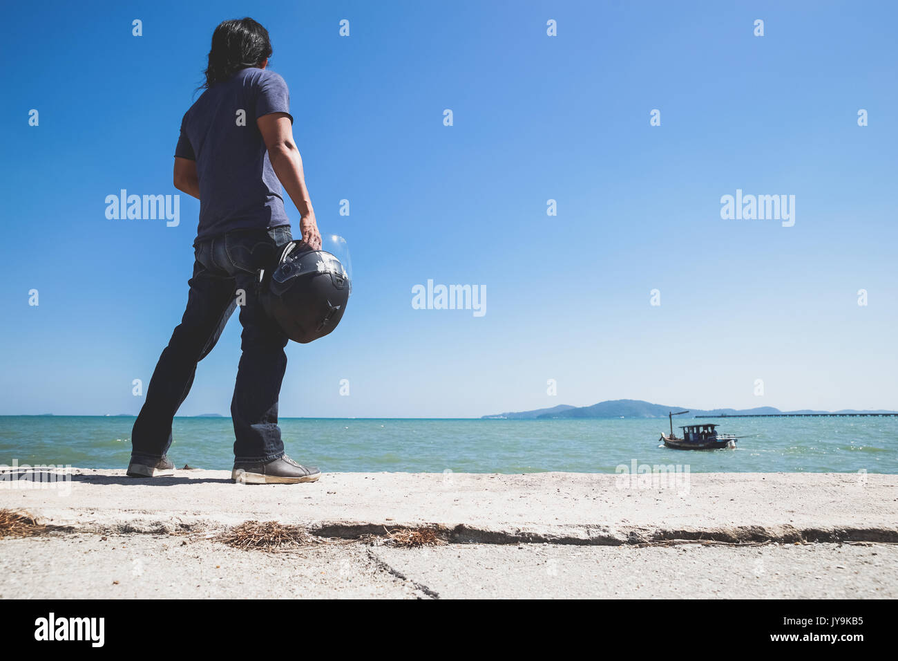 Un uomo asiatico stand girare la schiena accanto al mare mentre si tiene il suo casco e guardando alla barca Foto Stock