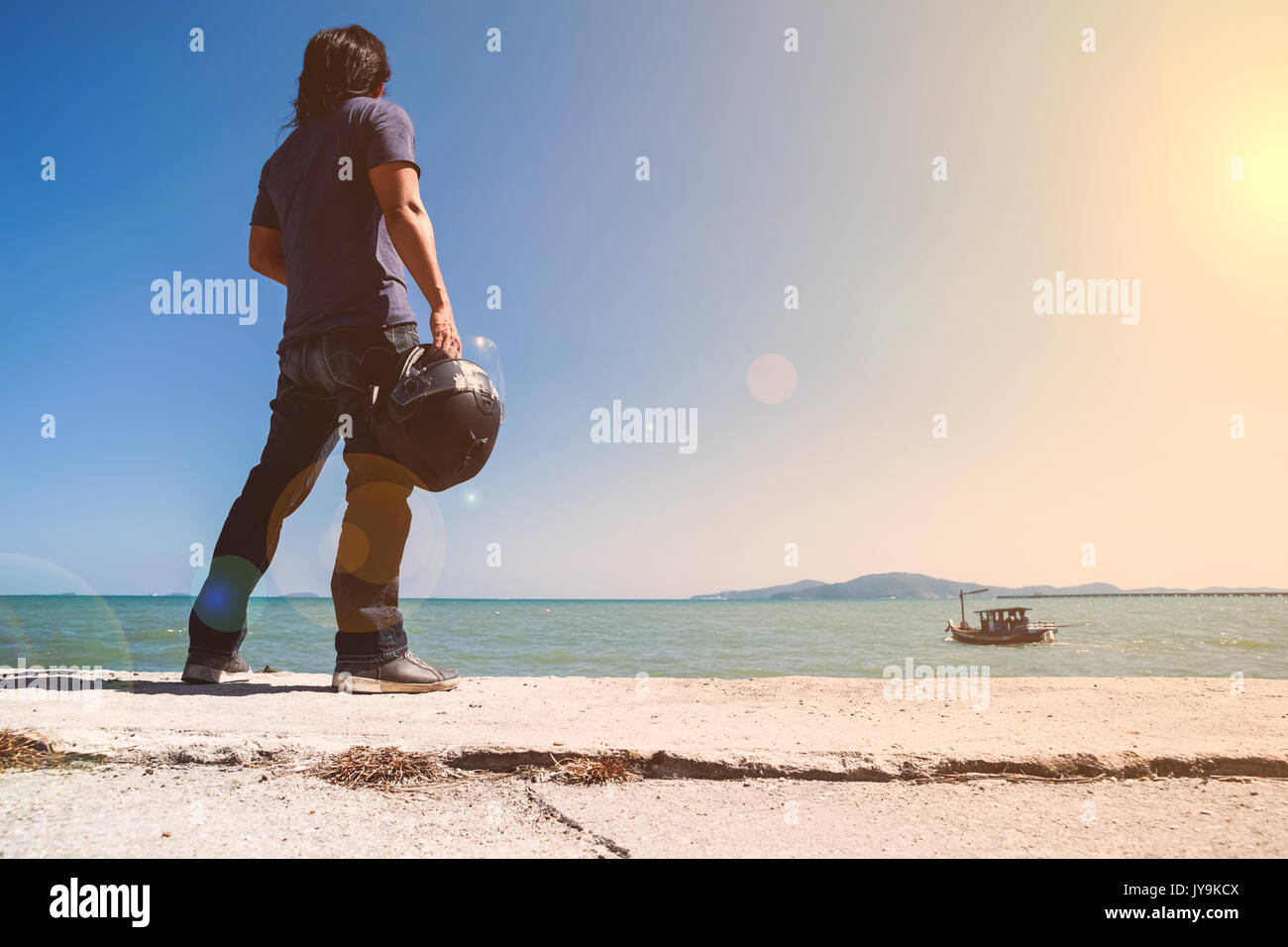 Un uomo asiatico stand girare la schiena accanto al mare mentre si tiene il suo casco e guardando alla barca Foto Stock