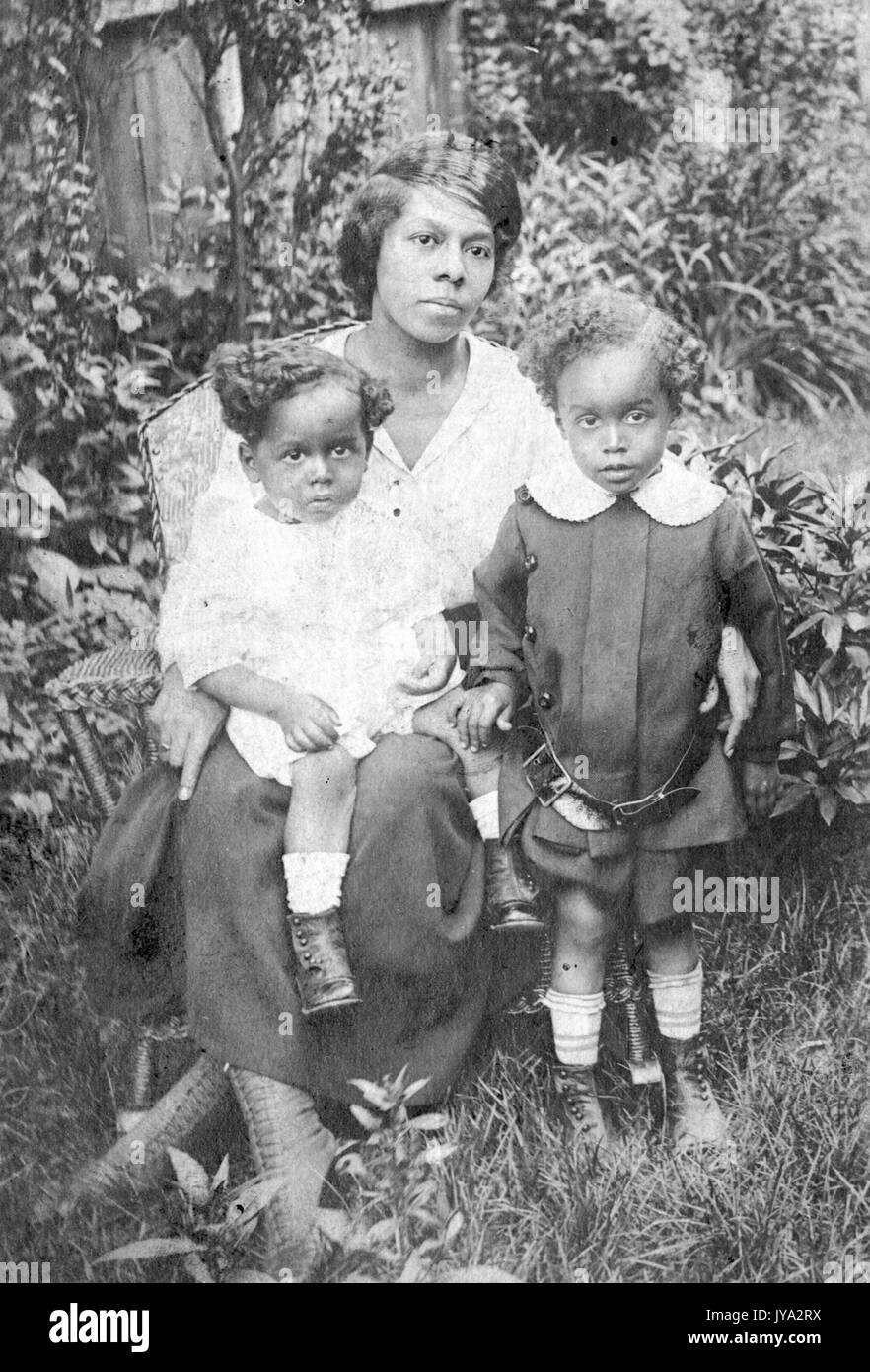 African-american madre e bambini in posa per una foto all'aperto, la madre seduto in una sedia di vimini, la sua giovane figlia seduta sul suo giro, suo figlio in piedi accanto a lei e indossando una cinghia allentata, tutti i vuoti con le espressioni del viso, bambini di età compresa tra 2 e 8, 1910. Foto Stock