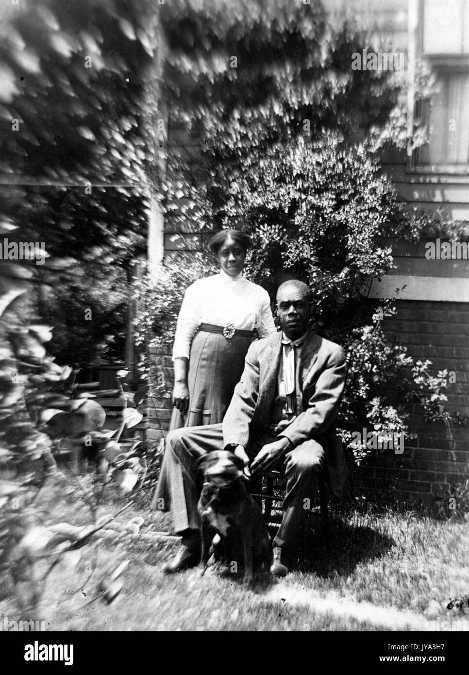 African American giovane, un uomo e una donna, seduto al di fuori della loro casa e in posa per fotografie con il loro cane, il movimento della telecamera creando l'effetto di movimento sul fogliame intorno al giovane, 1932. Foto Stock