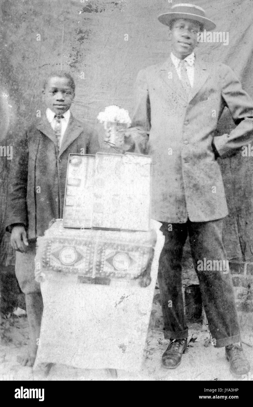 African American padre e figlio in posa per una fotografia, Padre indossa una tuta e in piedi con una mano sulla sua anca, il figlio che indossa un abito e cravatta, in piedi dietro un tavolo con puntelli, 1932. Foto Stock