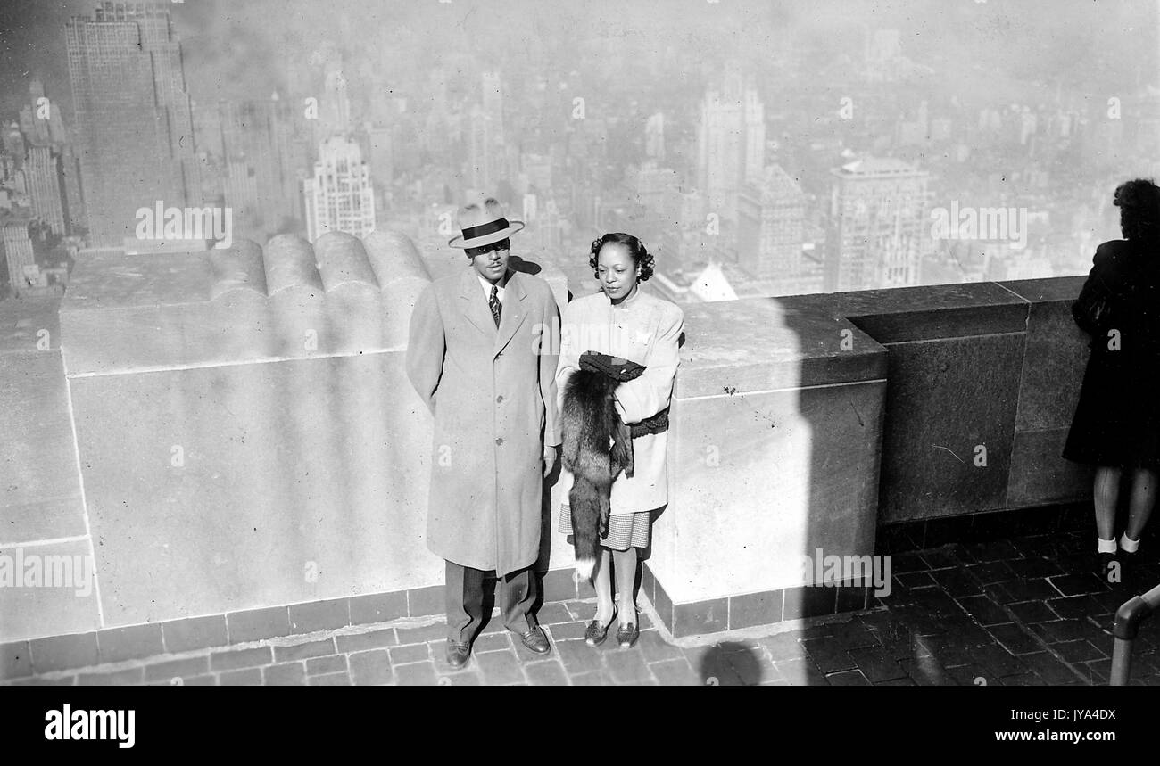 African-american giovane, un uomo e una donna, in piedi insieme vicino a una ringhiera in cima all'Empire State Building, su una piattaforma di osservazione, visto dal di sopra, lo skyline di New York visibile in background, l'uomo che indossa un trench coat e formale del cappello, la donna che indossa una maglietta e tenendo una pelliccia, 1946. Foto Stock