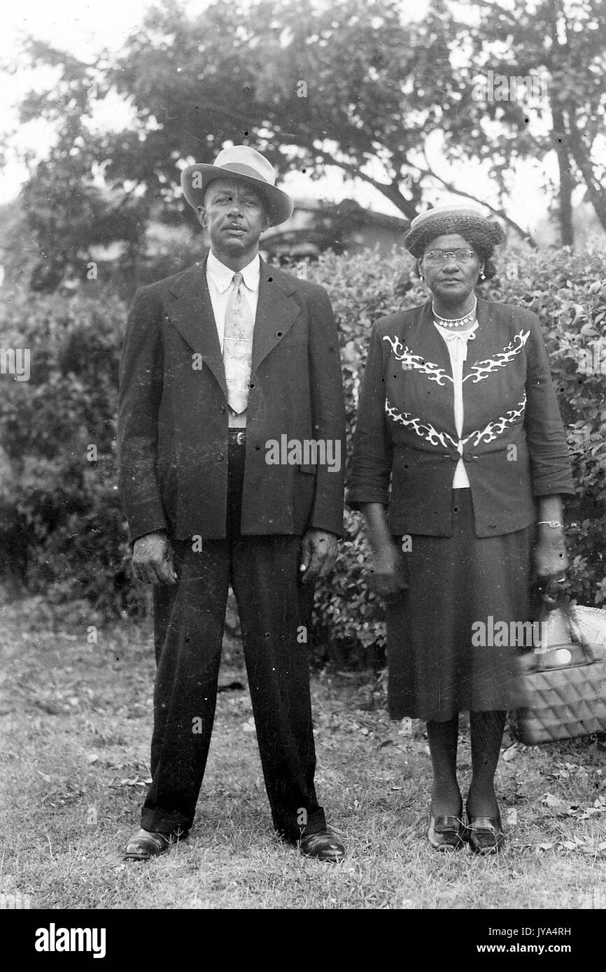 Coppia americano africano giovane in piedi insieme nel cortile di casa loro indossare abbigliamento formale, l'uomo che indossa un vestito e un cappello circolare, la donna che indossa un abito e tenendo una borsa, una siepe visibile in background, 1932. Foto Stock