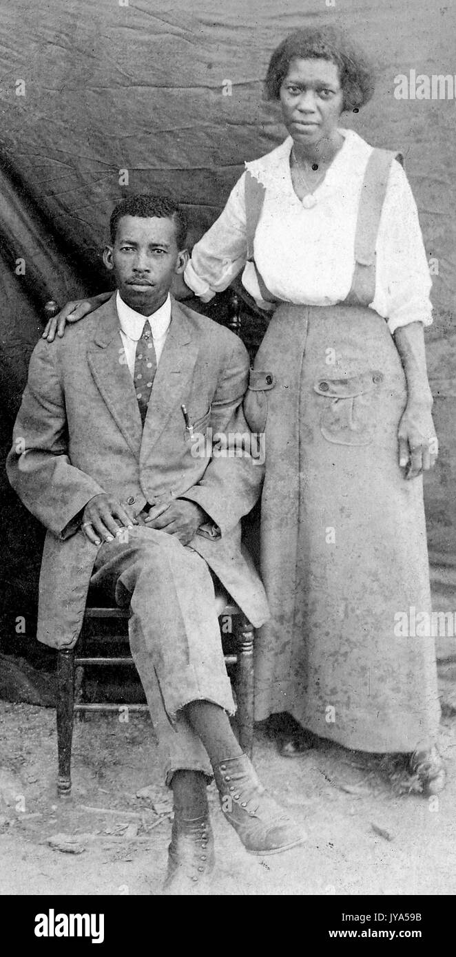African-american giovane in posa per una fotografia, tessuto di una tenda visibile in background, all'aperto, con la massa di polvere in una impostazione agricola, l'uomo seduto in una sedia, la donna in piedi e appoggiato la sua mano sulla sedia, indossando un abito usurato, 1912. Foto Stock