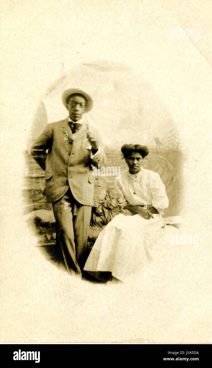 African-american giovane, un uomo e una donna, in posa per un ritratto in studio contro un fondale dipinto, l'uomo che indossa un vestito e un cappello di paglia e in piedi, la donna che indossa un abito bianco e seduto con le braccia incrociate in grembo, entrambi con gravi le espressioni facciali, 1905. Foto Stock