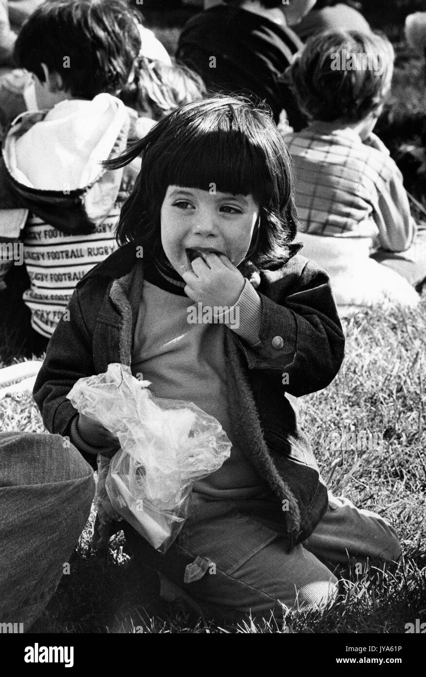 Una giovane ragazza si siede sull'erba mangiando cibo carnevale da un involucro di plastica, circondato da altri bambini e famiglie in background, durante il 3400 in Stage Festival alla Johns Hopkins University, Baltimora, Maryland, aprile 1978. Foto Stock