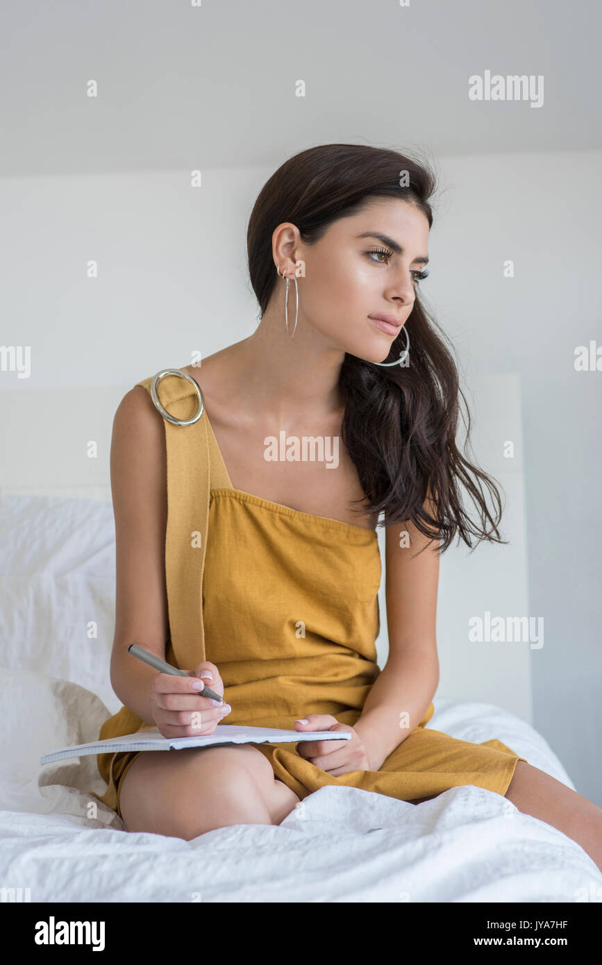 Giovane donna con lunghi capelli neri, seduta sul letto. Indossa un lungo abito marrone. Foto Stock