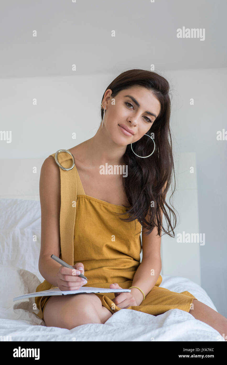 Giovane donna con lunghi capelli neri, seduta sul letto. Indossa un lungo abito marrone. Foto Stock