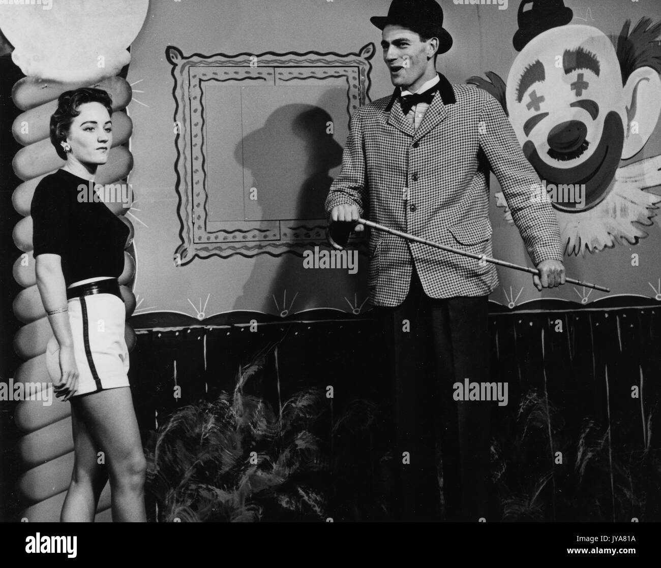 Due sono gli attori sul set riprese per la Johns Hopkins science review, attore lynn rosenthal è sulla sinistra e luce di indossare pantaloncini colorati e un nero manica corta shirt, attore edmond prelievo è a destra e indossa una tuta a scacchi giacca con pantaloni scuri, egli è in possesso di un magro di canna, dietro i due attori è dipinto di clown faccia su una parete, 1955. Foto Stock