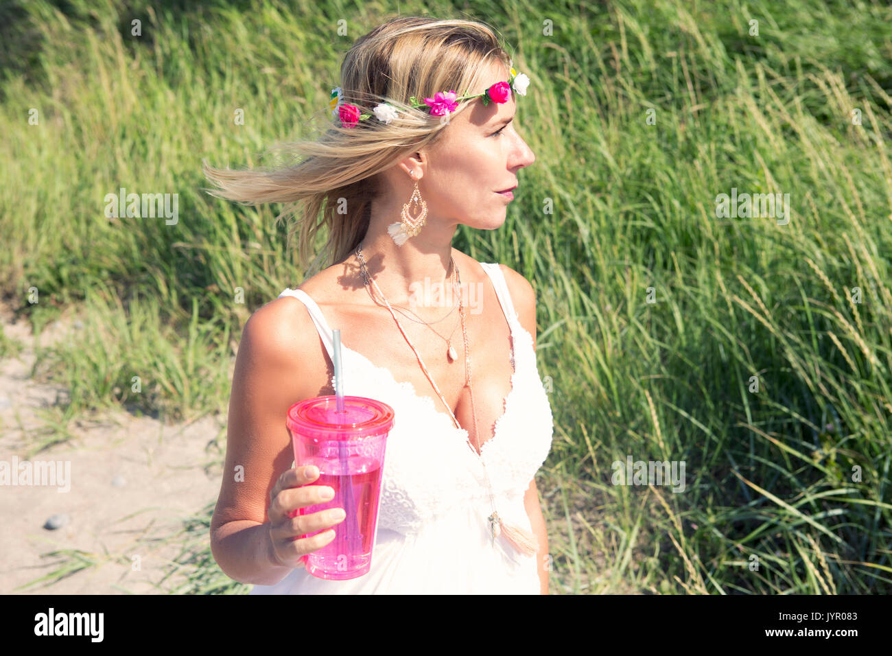 Donna bionda con fiori nei capelli e abito bianco permanente sulla spiaggia con bevanda Foto Stock