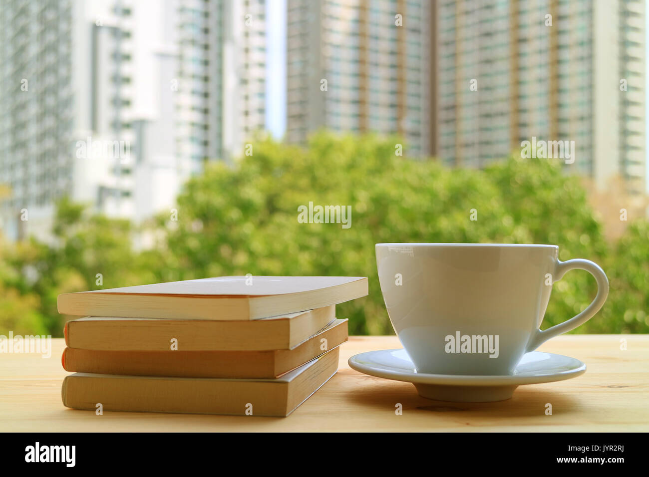 Rilassarsi con una tazza di una bevanda calda e alcuni libri dalla finestra, con sfondo sfocato di verdi alberi ed edifici alti Foto Stock