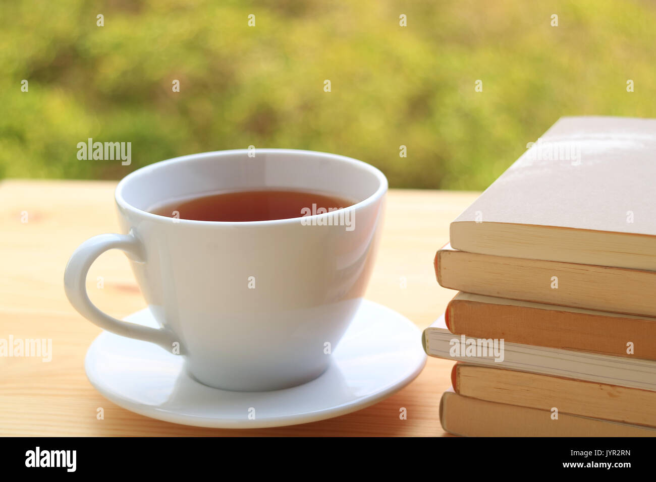 Tazza di tè caldo con la pila di libri sul tavolo di legno con sfondo sfocato di piante in giardino Foto Stock