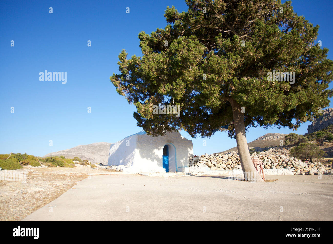 Agios Joanne, una piccola chiesa sulla Tilos Isola, Dodecaneso, Grecia sopra il porto di Livadia, abbandonato Gera ombreggiato sotto il grande albero di pino. Foto Stock