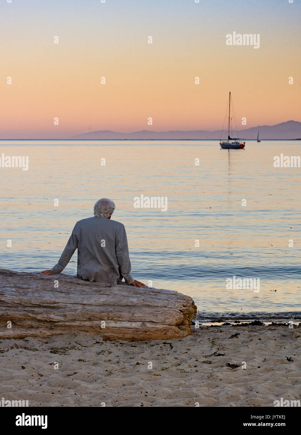 Grigio di un uomo dai capelli in una giacca beige seduto su driftwood guardando le barche a vela sull'oceano al tramonto. Foto Stock